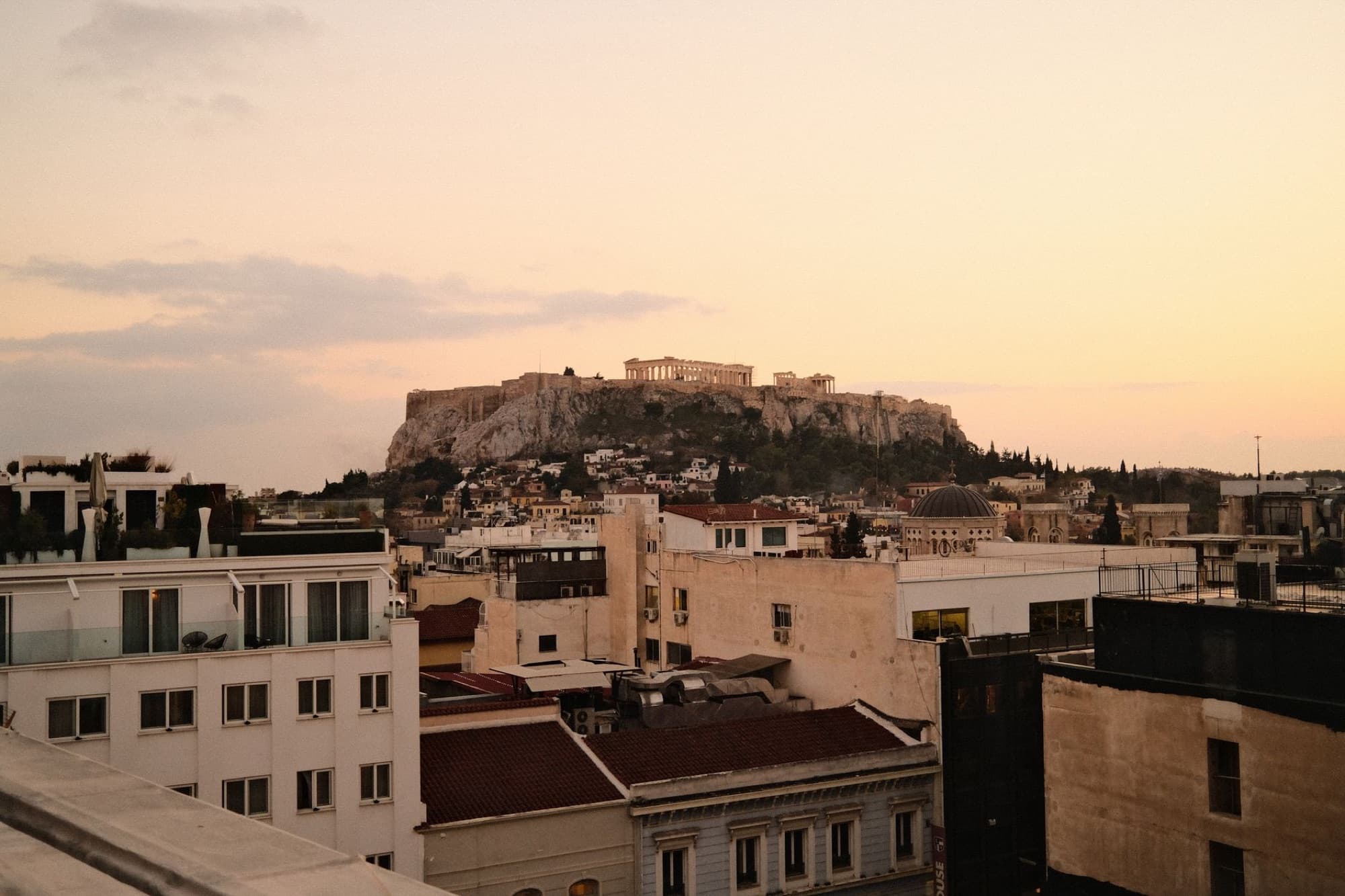 Scenic sunset view of the Acropolis in Athens, Greece, framed by city rooftops and a pastel sky.