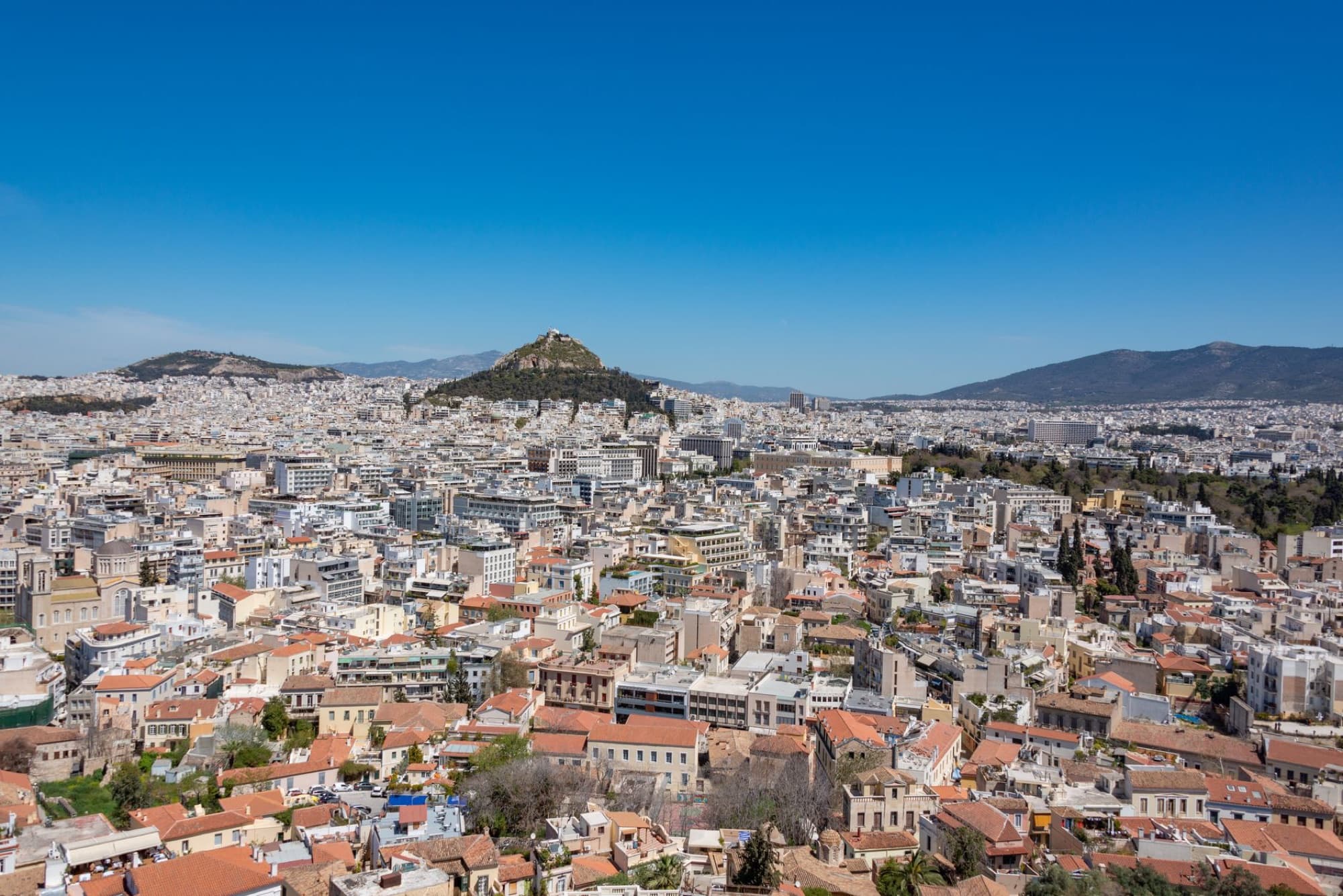 Expansive aerial view of Athens, showcasing the vibrant cityscape and iconic Lycabettus Hill under clear blue skies.