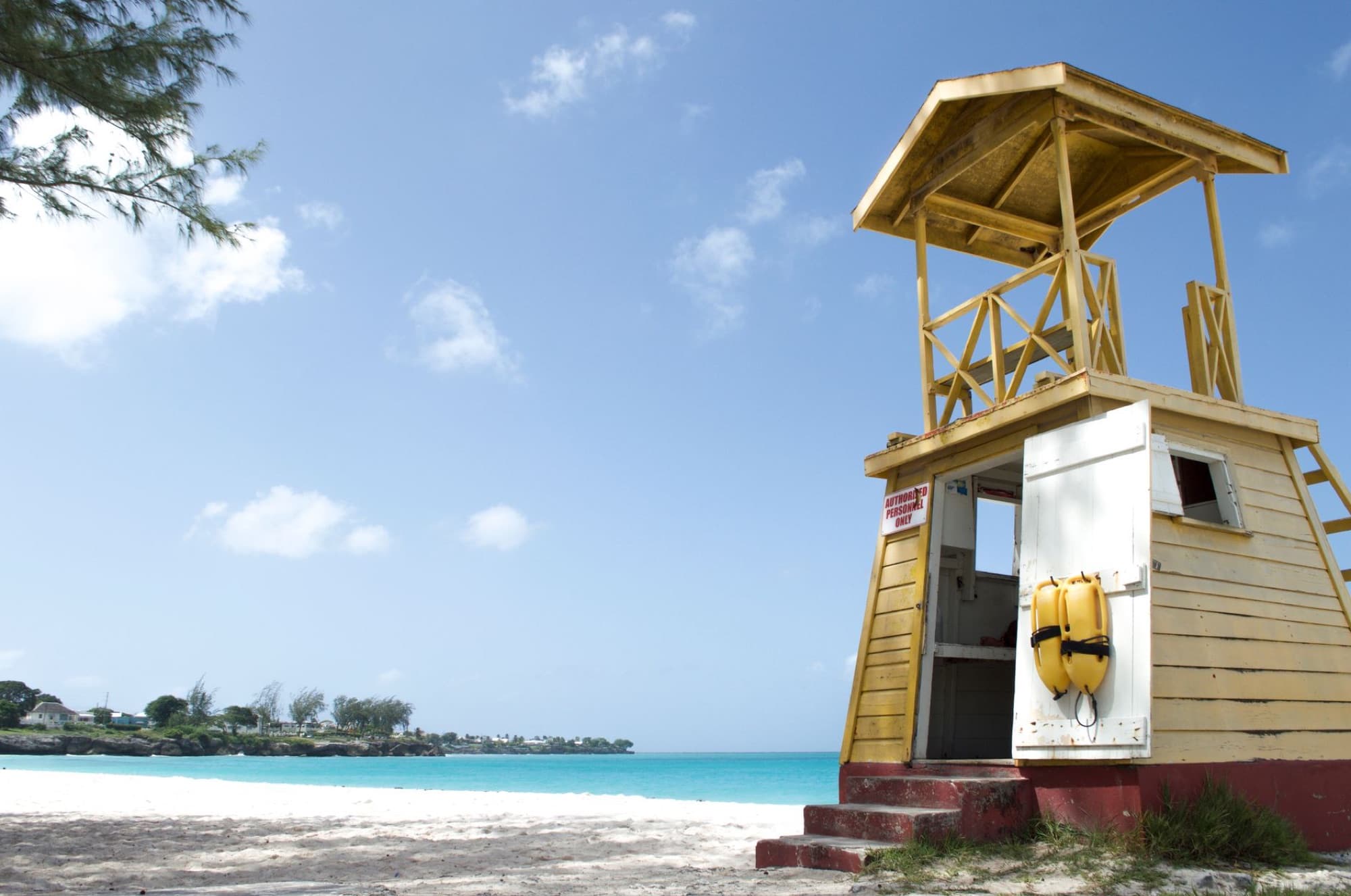 A wooden lifeguard tower stands on a sunny Barbados beach with clear blue skies and ocean.