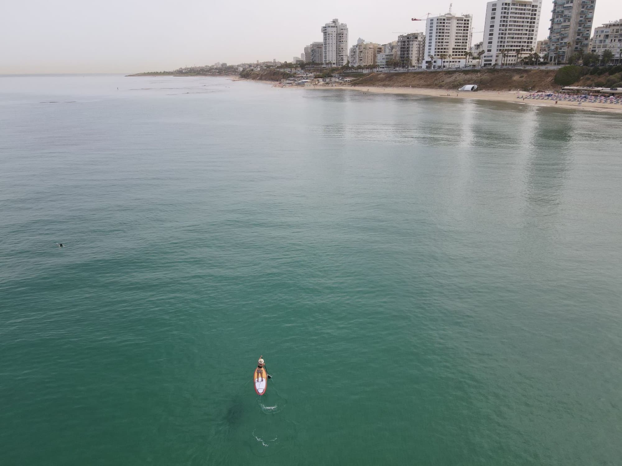 An aerial shot of a lone paddleboarder on calm sea near a city coastline.