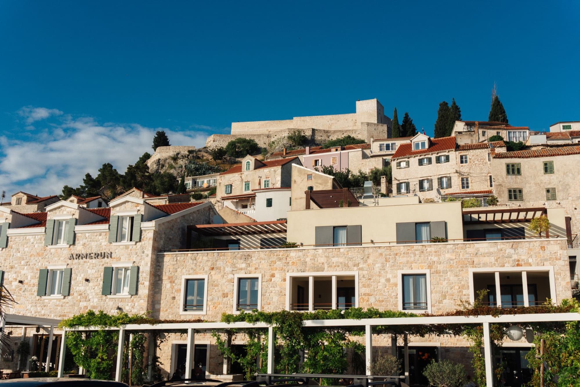 Scenic view of historic stone buildings in Hvar, Croatia with a fortress in the background under a clear blue sky.