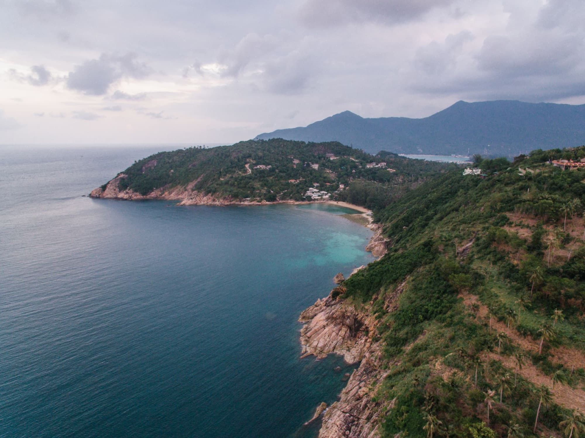 Breathtaking drone view of a coastal landscape with lush greenery, cliffs, and mountains.