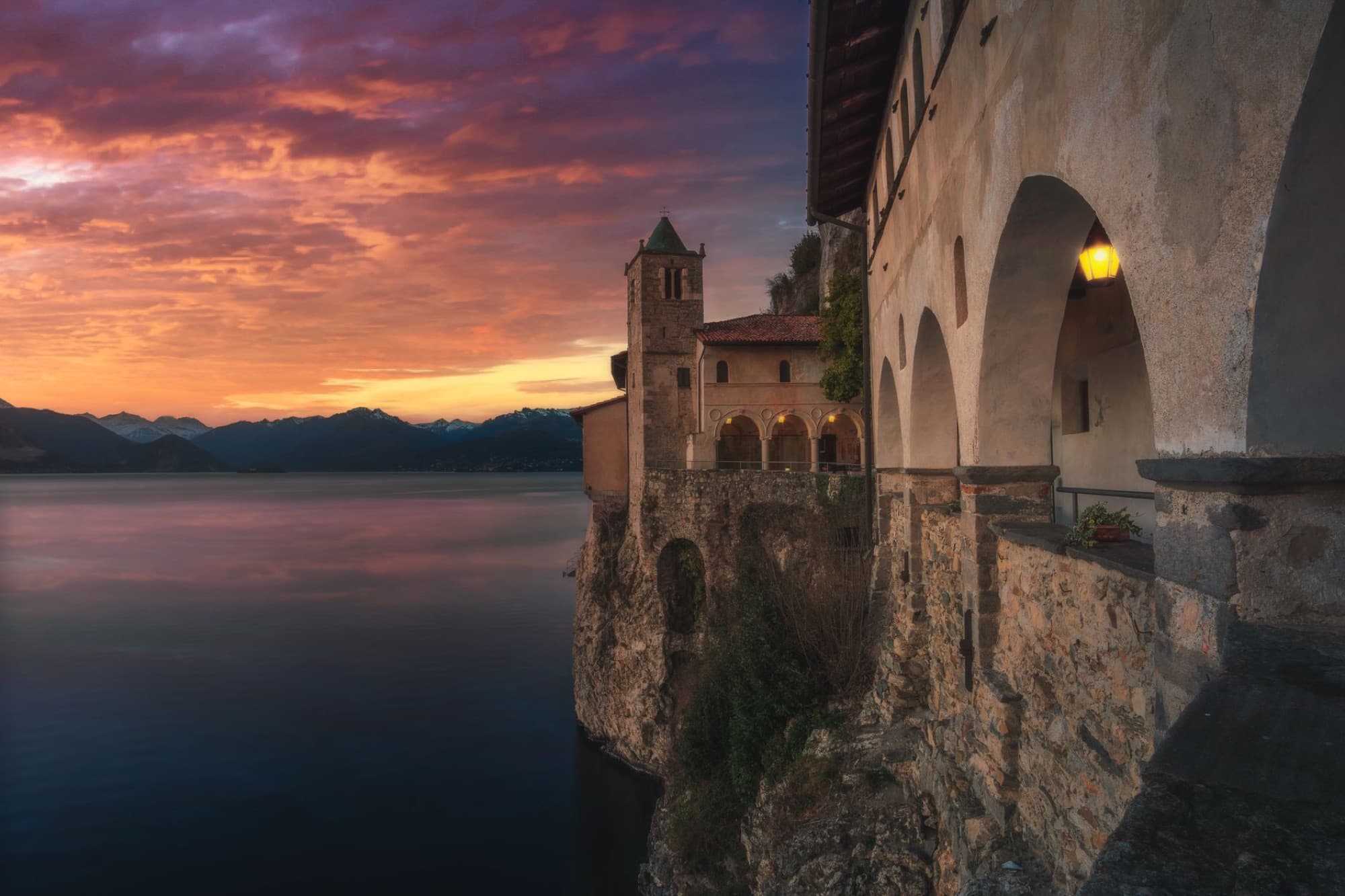 Scenic view of a historic monastery perched on a cliff overlooking Lake Maggiore at sunset in Italy.