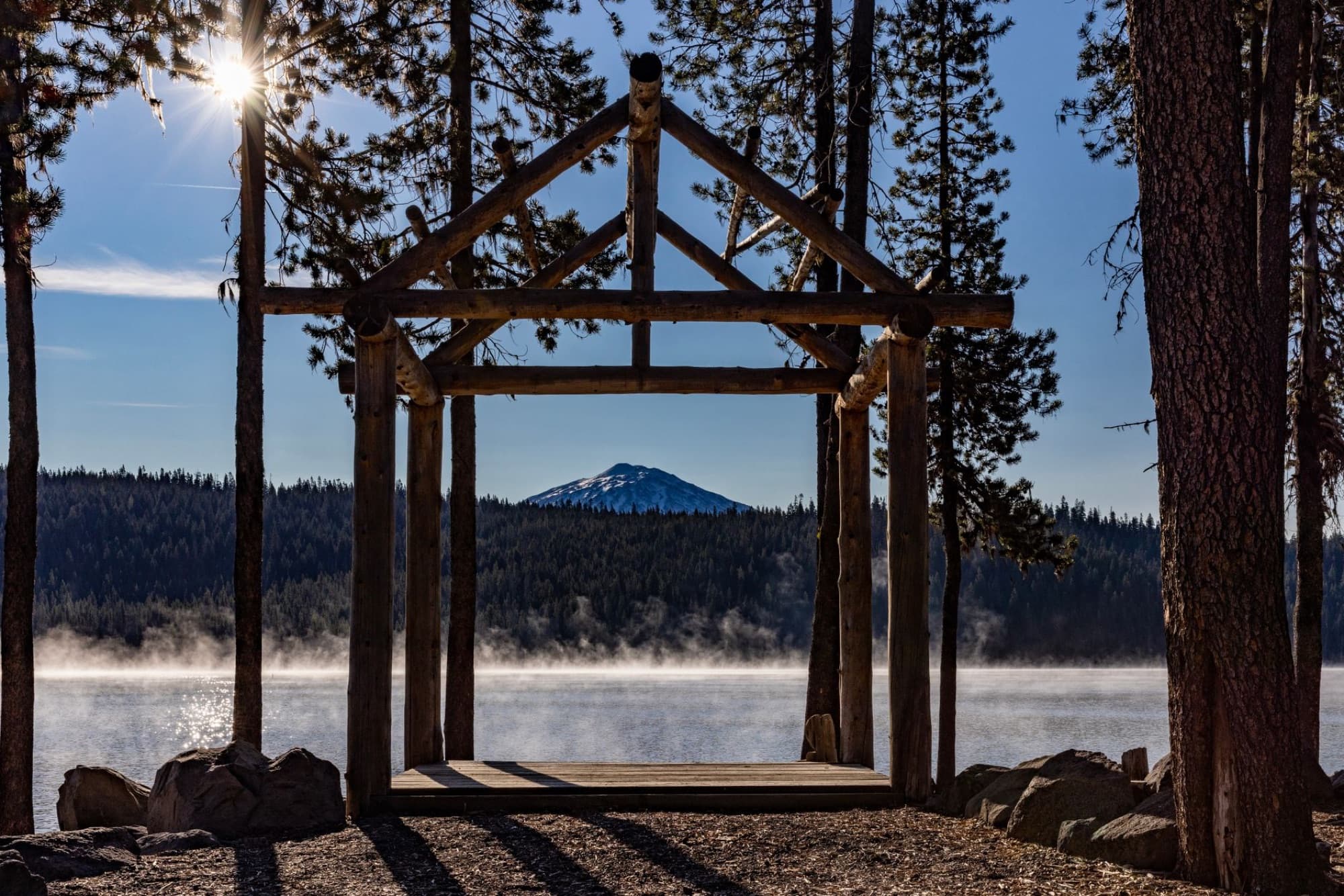 A rustic wooden structure framed by trees and sunlight with a scenic view of Elk Lake and distant mountain in Oregon.