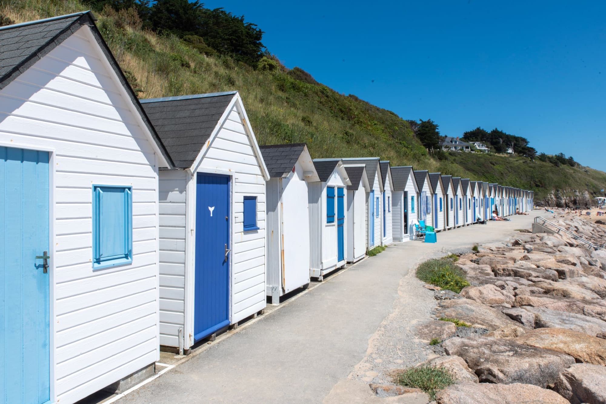 Row of white beach huts with blue doors along a scenic coastline under clear blue skies.