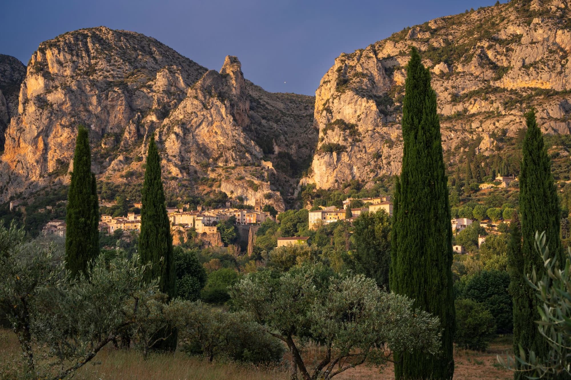 A scenic view of Moustiers-Sainte-Marie village with cypress trees and cliffs during golden hour in Provence, France.