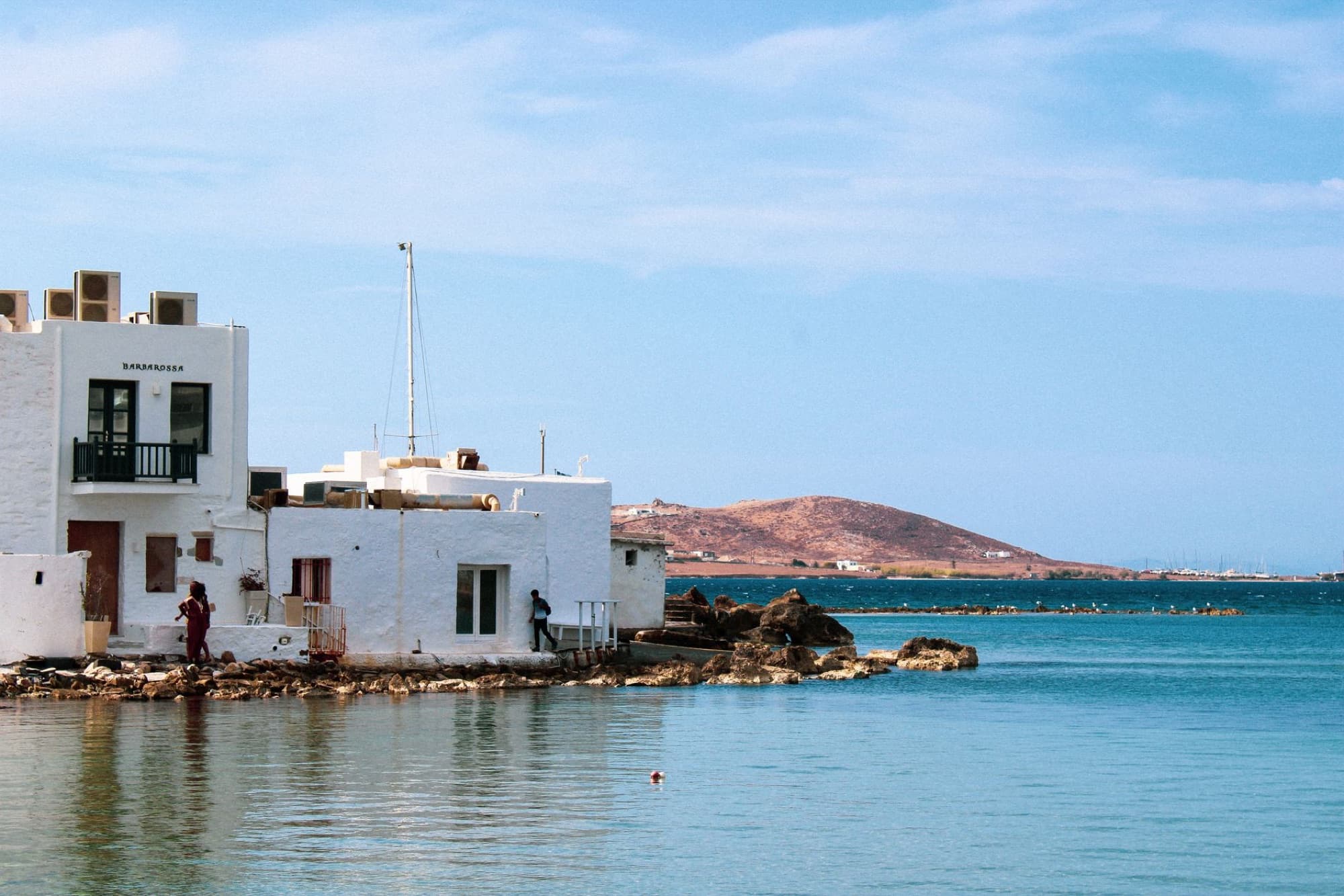 Charming white buildings by the coast in Naoussa, Paros Island with serene blue waters and hills in the background.