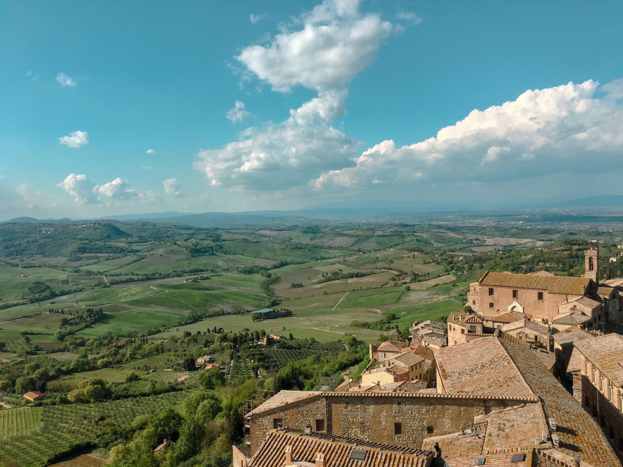 Aerial view of Montepulciano, Tuscany, featuring rolling hills, historic buildings, and lush greenery under a clear summer sky.