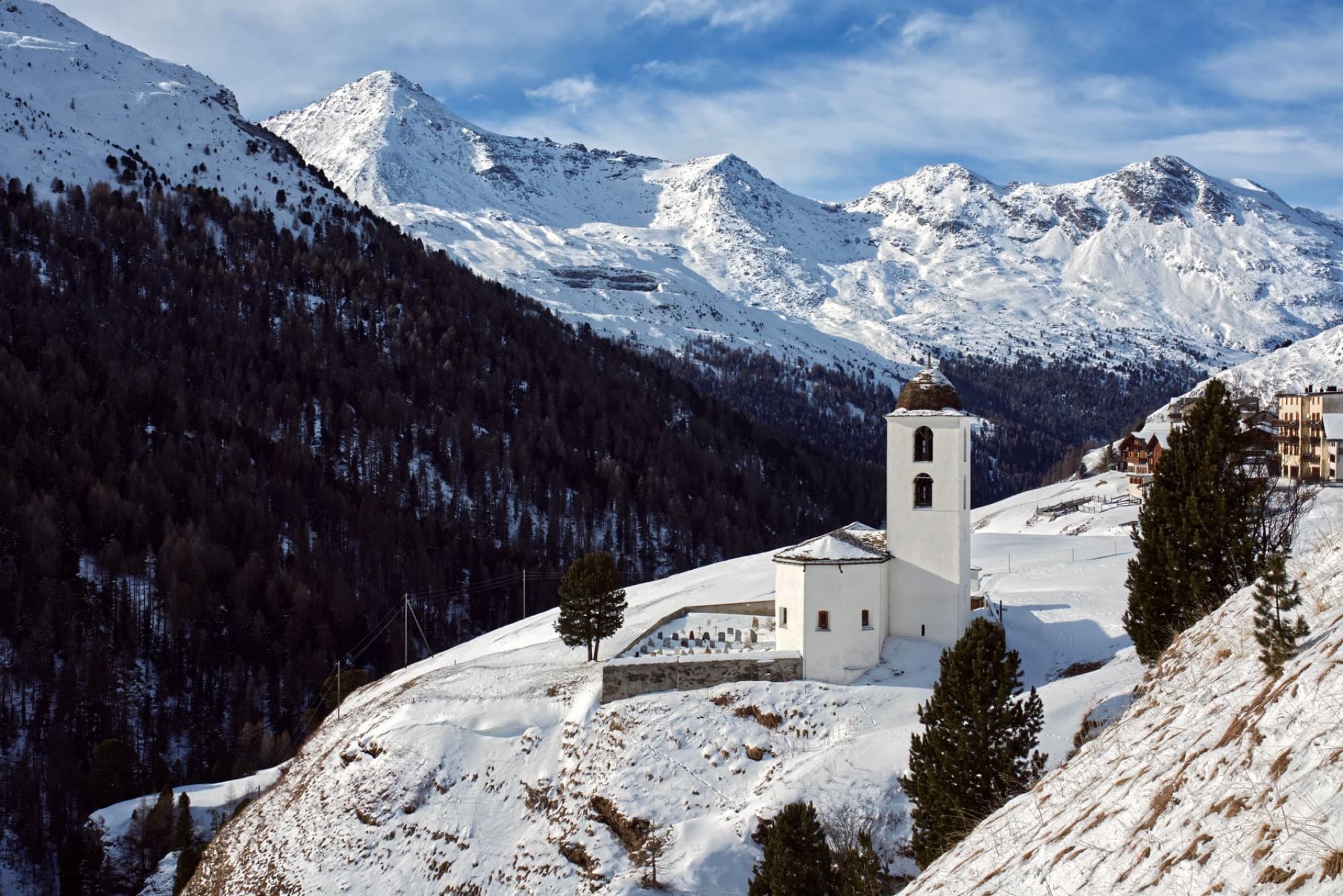A picturesque mountain chapel surrounded by snowy peaks and forest in a serene winter landscape.